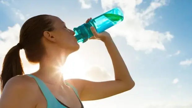 Ženska pije vodo iz steklenice, v ozadju sonce in modro nebo.