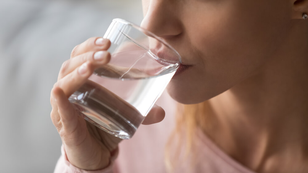 Ženska pije vodo iz kozarca, vidi se samo polovico njenega obraza in kozarec z roko